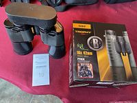 Bushnell Trophy 10x42 binoculars with box and MaxSold lot number card on red fabric surface.