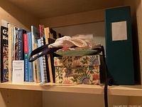 Shelf with assorted cookbooks and boxes, including 'Blitz Jones' Kitchen' and 'Cooking Light',' plus a cookbook holder and fabric items.
