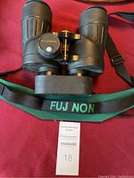 Top view of black Fujinon binoculars showing central focusing knob and eyepiece area with adjustments. Includes green strap marked 'FUJINON'.