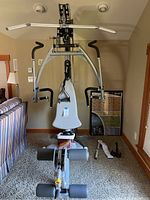 Front view of Hoist multi-station home gym showing overhead lat pulldown bar, chest press arms, padded seat and leg rollers