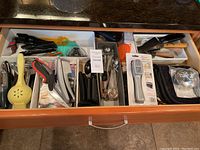Assorted kitchen utensils in a white drawer organizer, including juicers, can opener, garlic press, kitchen shears, spatula, slotted spoon, metal ladle, and various small tools