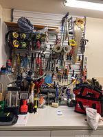 View of wall-mounted tool organizer filled with hand tools, measuring tapes, saws, hammers, and tool bags on and around workbench.