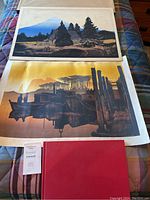 Two Elton Bennett serigraph prints and a red EB book laid out on a multicolored plaid fabric.