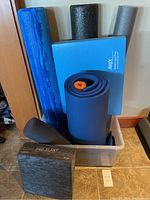 Full view of workout support items inside plastic bin showing foam rollers, wedge, blue Airex balance pad, and black Pro-Slant yoga block.