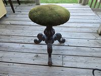 Antique piano stool with round green velvet seat and three carved wooden legs shown on a wooden deck floor.