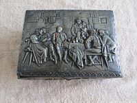 Top view of the closed rectangular metal box with repousse design showing a group of people around a table indoors with hanging laundry.
