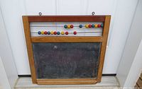 Frontal view of an antique slate chalkboard with wooden frame, featuring three rows of multi-colored counting beads mounted horizontally at the top. The slate surface shows wear and discoloration.