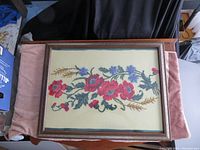 Top-down view of framed needlepoint with red poppies, blue cornflowers, green leaves, and wheat stalks on a cream background, inside a wooden frame with glass.