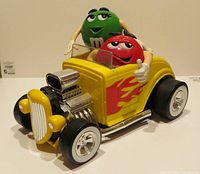 Yellow M&M Hot Rod candy dispenser with Red and Green M&M characters sitting inside, viewed from front and side showing flame decals and character details