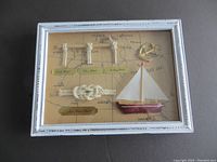 Front angled view of framed nautical wall decor. Shows white distressed frame, map background with knots, sailboat, and anchor.
