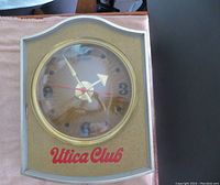 Front view of gold-textured and beige lighted clock with clear dome and red 'Utica Club' lettering on the bottom front panel.