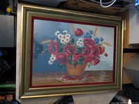 Full view of the framed needlepoint artwork showing a basket with red poppies, white daisies, blue wildflowers and wheat stalks on a wooden surface against a blue background.