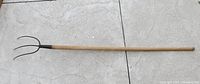 Full view of the 62 inch long vintage pitchfork with wooden handle placed on a concrete floor.
