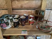 Eight assorted Christmas ceramic mugs, matching small plates, and a large hand-painted glass bowl displayed on wooden shelving.