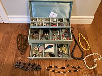 Open view showing various compartments filled with assorted costume jewelry pieces including necklaces, earrings, brooches, and bracelets, laid out inside a light blue jewelry box.