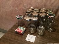 20 assorted Mason jars with various metal lids arranged on a table including decorative grape design lids and a box of Bernardin Mason jar bands.
