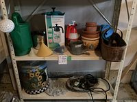 Wide shot of shelf containing green watering jug, sunflower storage can, garden tools basket, clay pots, sprayers, and funnel