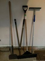 Four garden and cleaning tools standing against a wall: snow shovel, ice chipper, edging shears, and squeegee