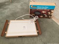 Full view of the vintage rectangular electric crepe maker on a wooden tray with electrical cord and original box in the background.