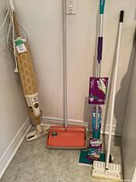 Photo shows a vintage Regina Elektrikbroom upright vacuum with beige and gold fabric bag, plugged in; orange vintage Bissell 400 carpet sweeper; Swiffer WetJet mop and a white mop all standing against a wall on tiled floor.