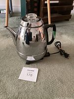 Full view of the chrome electric coffee percolator on the floor with power cord and sticker.