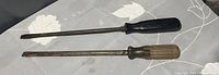 Two large screwdrivers lying on a surface, one with a black handle and the other with a brown wooden handle.