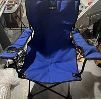 Blue adult folding camping chair with black metal frame, padded armrests, and cup holders on each armrest. Chair is folded open and placed on a basement floor.