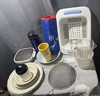 Photo showing assorted kitchen items on a table including plates, colander, measuring cups, strainer, grater, drinkware, and an insulated bag.