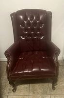Front view of a burgundy vinyl accent chair with button-tufted back and seat, wingback style with nailhead trim on arms and cabriole legs, placed on tiled floor against white wall.