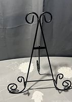 Black metal large picture frame stand with curved ornamental accents at base and top, shown on a fabric surface with a neutral background.