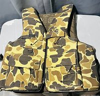 Front view of camouflage fishing/hunting vest with multiple pockets, zipper, and D-ring detail.