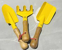 Three mini yellow metal garden tools with wooden handles: trowel, rake, and scoop-shaped shovel placed on a gray surface.