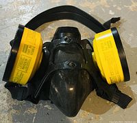 Front view of black silicone respirator mask showing the two attached yellow cartridge filters and elastic straps.