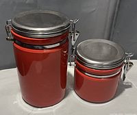 Photo showing two red ceramic canisters with black lids secured by metal clasps on a neutral background.