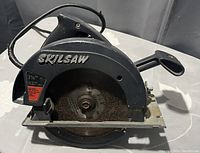 Black Skilsaw 7 1/4 inch circular saw on white surface, showing blade and handle.