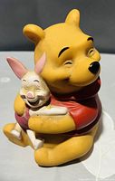 Photo of a vintage Winnie the Pooh and Piglet figurine showing Pooh hugging Piglet while both smile, captures color and condition.