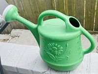 Green plastic watering can with floral embossing and white spray nozzle shown on a white brick surface outdoors.