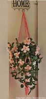 Full view of the pink silk flower plant hanging from a wall hook with the crocheted pink hanger attached to a white ceramic pot.
