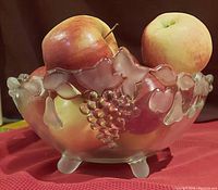 Front side view of the brushed glass fruit bowl filled with apples, showing grape cluster and leaf design around scalloped rim and three of the four legs.