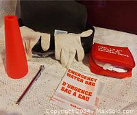 Photo of emergency car kit items including red first aid kit pouch, white cotton gloves, small fluorescent orange cone, and emergency water bag on white lace surface.