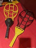 Two mini lacrosse paddles, one yellow and one black, on red fabric background taken from an angle showing full length.