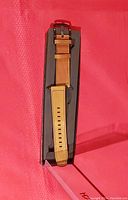 Soft tan leather watchband displayed inside an open black box, showing the entire band with buckled end and adjustment holes.