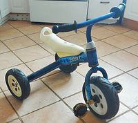 Front-side angle of royal blue tricycle with white banana style seat, black rubber wheels with white hubs, and black handle grips. Pedals visible at front wheel.