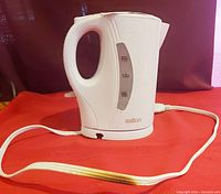 White Salton cordless kettle front view showing water level window, handle, and cord with plug on red surface