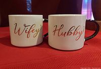Front view showing gold script 'Wifey' and 'Hubby' on white porcelain mugs with black handles.