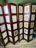 Two Umbra tall wood panel room dividers with photo frames, folded and standing side by side against a green wall.