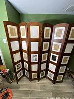 Two tall wooden Umbra room divider screens, each with three hinged vertical panels containing multiple built-in photo frames holding placeholder images. The wood is warm-toned and smooth with slightly curved tops on panels.