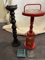 Photo showing two standing ashtrays and two tabletop ashtrays on floor: tall dark wood turned ashtray with metal insert, red hand-painted standing ashtray with crystal glass inset, blue ceramic ashtray and clear glass ashtray.