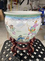Front view of ceramic planter showing the hand painted scene of children playing and a pagoda, with the wooden stand below.