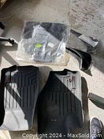 Two black WeatherTech floor mats, one still in plastic packaging, two black Dodge mud guards, all laid on concrete floor with some shadows.